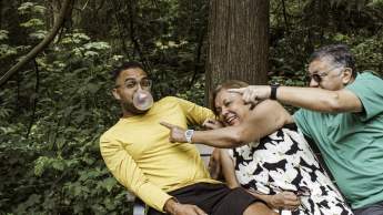 A man leaning on a bench popping bubblegum with an elderly couple trying to pop the bubble.