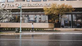 A person walks past The Coast Bastion Inn on a rainy day, with wet pavement and trees lining the street.