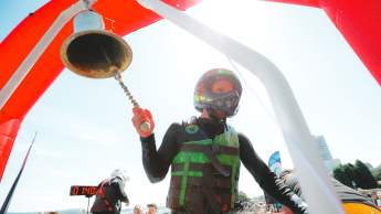 A racer wearing a helmet and life jacket rings a finish bell beneath a red arch as a crowd cheers along the waterfront behind him.
