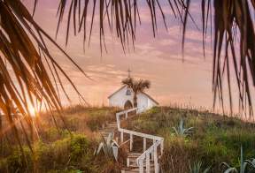 On top of a grassy hill at sunset sits a small white chapel topped with a cross. Steps with a white railing lead up to the chapel and palm tree fronds frame the photo