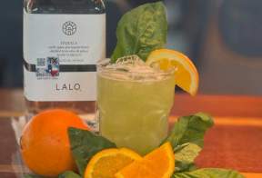 A light green margarita sits on a table surrounded by oranges and basils
