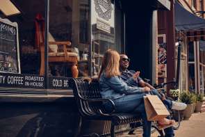 Two people sitting on a bench with a shopping bag, in front of a coffee house.