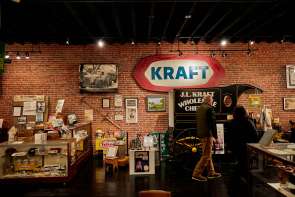 Two people are touring a museum with signs from the past, like Kraft, with a brick wall behind them.