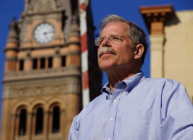 John Gurda stands in front of Milwaukee City Hall, looking into the distance with the landmark's iconic clock tower rising behind him. The close-up portrait is set against a clear blue sky, highlighting one of Milwaukee's most recognizable architectural icons.