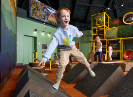 A child excitedly balances on angled foam blocks in the “Hot Lava” obstacle course exhibit at the Betty Brinn Children’s Museum, with other children playing in the background.