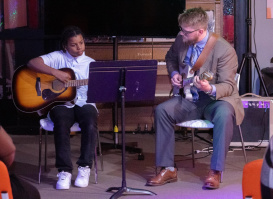 Two people seated and playing acoustic guitars during a live indoor performance.