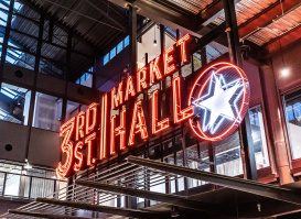 Neon sign reading ‘3rd Street Market Hall’ with a glowing star, inside a modern industrial-style building with glass walls and metal beams.