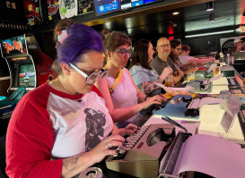 A group of people sit side by side at a bar counter, each using a typewriter. The person in the foreground has purple hair tied back with a scrunchie, glasses, and a red-and-white shirt with a graphic print. Others focus intently on their typewriters, with drinks and cans on the counter. Behind them, arcade machines, merchandise, and a TV screen are visible, creating a lively retro atmosphere.