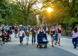 Visitors stroll through the Milwaukee County Zoo during a summer evening event, with the sun setting behind trees and a fountain in the background.