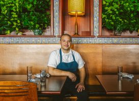 A chef wearing a white shirt and blue apron sits at a wooden booth inside a restaurant, smiling at the camera. The background features leafy green plants, patterned tile accents, and a warm lamp centered above him.