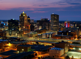 A twilight view of downtown Milwaukee, Wisconsin, featuring illuminated skyscrapers and city lights against a purple and blue sunset sky, with highways and streets visible in the foreground.