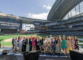 Group photo of attendees at Visit Milwaukee's Annual Meeting, posed on a stage at American Family Field (Milwaukee Brewers stadium) with stadium seating and banners visible in the background.