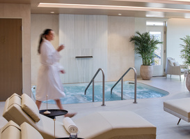A tranquil spa lounge features cushioned reclining chairs, rolled towels, and a bubbling hydrotherapy pool beneath warm wood-paneled ceilings. A guest in a robe walks through the bright, modern space, creating a relaxing atmosphere focused on wellness and rejuvenation.