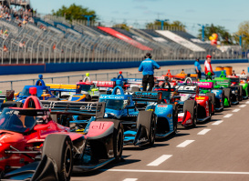 IndyCar race cars lined up on the track at the Milwaukee Mile with drivers and officials preparing for the start of the race, as spectators watch from the grandstands under a sunny sky.