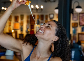 A smiling woman pulls apart a cheesy appetizer, creating a long stretch of melted cheese as she enjoys a meal inside a warmly lit restaurant. The background is softly blurred with artwork and lights visible.