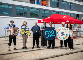 A welcoming committee with an accordion player stands before a cruise ship, holding Milwaukee-themed signs including an MKE logo and "City of Festivals" circular display, under a red Viking Cruises tent.
