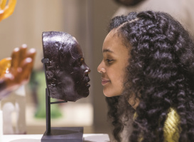 A young visitor leans in to closely examine a glass face sculpture displayed on a stand at a museum. The side profiles of the sculpture and the visitor align, creating a striking visual comparison in the gallery setting.