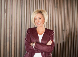A woman with short blond hair stands smiling with her arms crossed in front of a wall of vertical wooden slats. She wears a burgundy leather jacket over a white top and black pants.