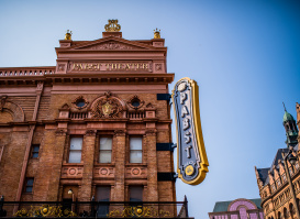 The exterior of the historic Pabst Theater in Milwaukee, featuring its ornate brick façade, gold architectural details, and large vertical “Pabst Theater” marquee sign against a clear blue sky.