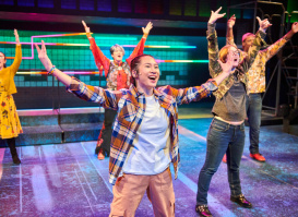 A group of young performers on a brightly lit stage throw their arms up mid-dance, smiling and full of energy, with colorful set pieces glowing behind them.