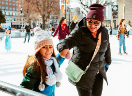 a little girl ice skating on a public ice rink while holding her mom's hand
