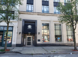 Street view of a beige stone building with tall windows and two trees on the sidewalk. The entrance has a black awning with a white “G” logo and the word “GOODKIND” above the door, along with the address number 333.
