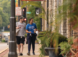 Two people stroll along a lively downtown sidewalk lined with local shops, hanging business signs, and colorful planters. Decorative lights and welcome banners add to the inviting streetscape as they explore the district on a warm day.