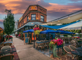 A brick building set against a sky at dusk, with orange and blue tones, a patio lit up with string lights, blue umbrellas and pink and yellow flowers.