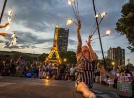 A fire performer in a striped costume kneels and raises flaming batons during a lively Bastille Days event in Milwaukee, with a crowd watching and a glowing Eiffel Tower replica lit up in the background.