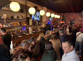 A crowded bar with warm lighting and exposed brick walls. People are gathered closely together, talking and drinking. A long bar counter is lined with bottles and glassware, and several round hanging lights illuminate the space. Two TVs mounted on the back wall display a show, and the overall atmosphere appears lively and social.