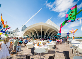 A wide angle shot of the Milwaukee Art Museum with festival goers sitting on the patio outside, with flags blowing in the wind against a beautiful blue sky.