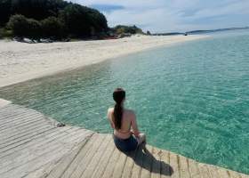 Woman sat on jetty overlooking turquoise waters and a sandy beach