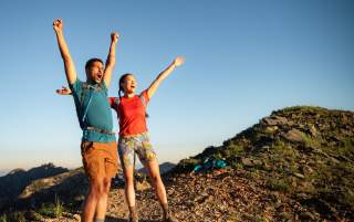 Man and Woman wearing hiking gear with their arms up and looking excited to reach the top of a mountain