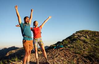 Man and Woman wearing hiking gear with their arms up and looking excited to reach the top of a mountain