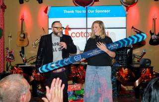 Two people are standing in front of a crowd. One holds a microphone the other is holding dynastar skis