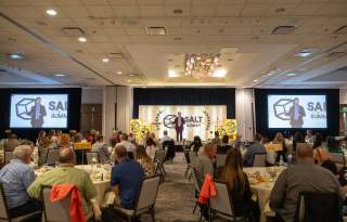 Man in brown suit speaking on a stage with the logo of the SALT Summit behind him and round tables with people attentively listening
