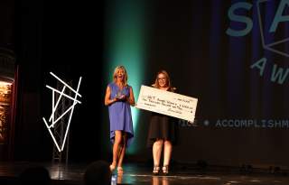 Two women standing on stage. One holds a big check made out the the Salt Awards winner. The other is holding a crystal award statue.