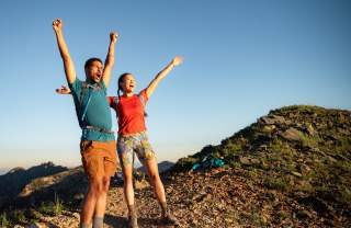 Man and Woman wearing hiking gear with their arms up and looking excited to reach the top of a mountain
