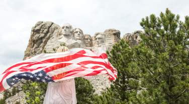 A woman holding an American flag behind her faces away from the camera toward Mount Rushmore, visible in the upper half of the photo under a mostly cloudy sky.