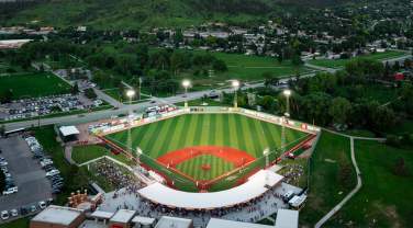 A high-angle view of Fitzgerald Stadium from behind home plate at dusk. The stadium lights illuminate the playing surface as the dusk sky darkens. The hills beyond center field are a vibrant green in peak summer.
