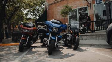 Three motorcycles, one red, one blue, and one black, are parked along the sidewalk, partially shaded by a tree, outside of Prairie Edge in Downtown Rapid City in mid afternoon.