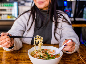 woman smiling while using chopsticks to eat a bowl of ramen