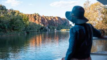 A woman enjoying the scenery on an Ord River cruise - the woman is standing on the vessel looking across the water at a cliff faced bathed in sunlight.
