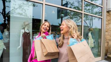 A laughing daughter with blonde hair looks over to her smiling mother as they carry shopping bags during a shopping spree in Overland Park.
