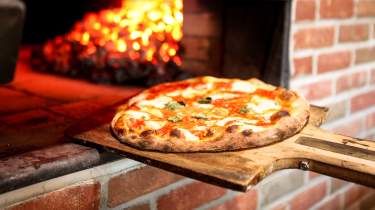 A perfectly cooked pizza is being pulled out of a woodfire oven on a wooden paddle.