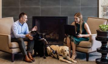Two people are seated in front of a fireplace discussing a plan for a presentation. One person has a laptop and two service dogs sit on the floor between the people.