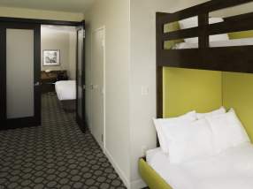 Family suite at Hyatt Regency Orange County featuring a bedroom area with a bright yellow headboard, white bedding, patterned carpet, and an adjoining living space visible through an open doorway.