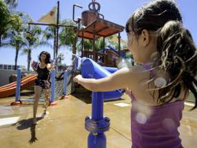 Howard Johnson by Wyndham Anaheim Hotel and Water Playground featuring a child using a water cannon at an outdoor splash pad, with play structures, slides, and another person in the background under a sunny sky.