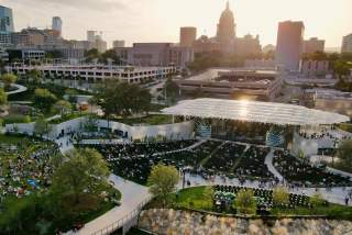 Aerial view of Moody Amphitheater in evening sun. There is a crowd on the open lawn and in seats near the pavilion stage. The State Capitol building is visible behind the amphitheater