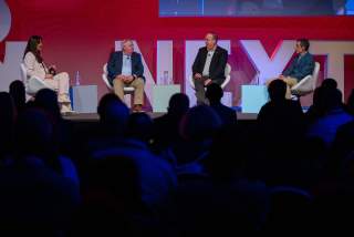 A woman sitting on a stage with three men to her right, speaking on a panel in front of a dark room of attendees.